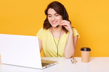 Kulaklık gülümseyen neşeli destek telefon operatörü kadın Stüdyo çekim, sarı arka plan üzerinde izole müşterisi ile sohbet etti, hoş yüz ifadesi vardır, beyaz masada oturan.