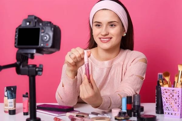 Imagen de una joven adorable prueba algunos productos de belleza y muestra brillo de labios a sus seguidores en el blog. Belleza vlogger se sienta delante de la cámara rodeada de cosméticos, tiene expresión agradable . — Foto de Stock