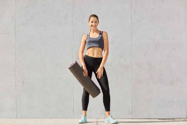 Atractiva joven deportista hermosa con top, leggins y zapatillas de deporte, con cola de caballo, celebración de estera de yoga, tiene entrenamiento, entrenamiento interior, aislado sobre fondo gris. Concepto de salud . —  Fotos de Stock