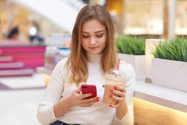 Beautiful blonde student sitting in cafe, reading text message on her smart phone and drinking lemonade from takeaway plastic cup, dressed casually, comunicates with friend, uses wireless Internet. — Stock Photo, Image