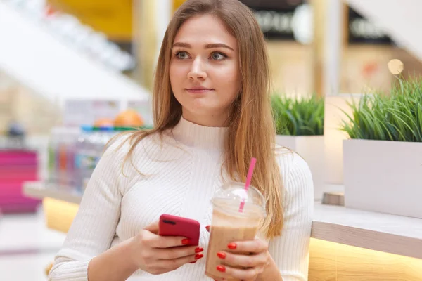 Portrait of beautiful girl, has European appearance, dressed white sweater, sits in cafe, drinks lemonade, holds telephone in hand, looking aside, talks with friend. People and spare time concept. — Stock Photo, Image