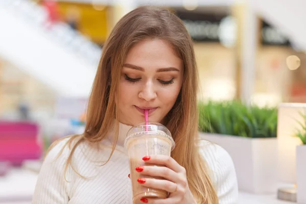 Фото чарівного блондинки-жінки п'є коктейль, позує в кафе, носить білий повсякденний светр, має червоний манікюр, дивиться вниз на її напій, любить його смак, насолоджується виходами, концепцією людей та молоді . — стокове фото