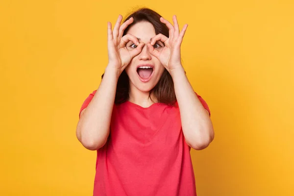 Εικόνα της νεαρής ευτυχισμένη γυναίκα με κόκκινο μπλουζάκι, κρατώντας το χέρι της πάνω από τα μάτια της ως γυαλιά και κοιτάζοντας μέσα από τα δάχτυλα, διασκεδόντας ενώ φωτογραφίζονται σε στούντιο, απομονώνονται σε κίτρινο φόντο. — Φωτογραφία Αρχείου
