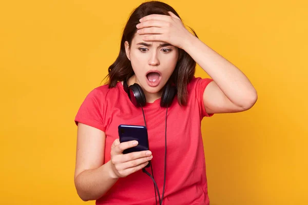 Close up portrait of stupefied Caucasian woman opens mouth widely, reads incredible news in social network, holds cell phone, dressed in red casual t shirt, poses indoor isolated on yellow background. — Stock Photo, Image