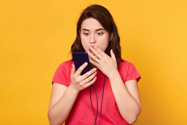 Close up portrait of shocked woman talking to somebody, has video call, uses modern smartphone, dressed red t shirt, covers mouth with hand, hears surprised news, has astonish facial expression. — Stock Photo, Image