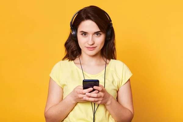 Shot of young dark haired girl with earphones, ready to listen to music, looks derictly at camera with calm facial expression, wear yellow t shirt, spend free time alone, isolated on studio wall . — Stok Foto