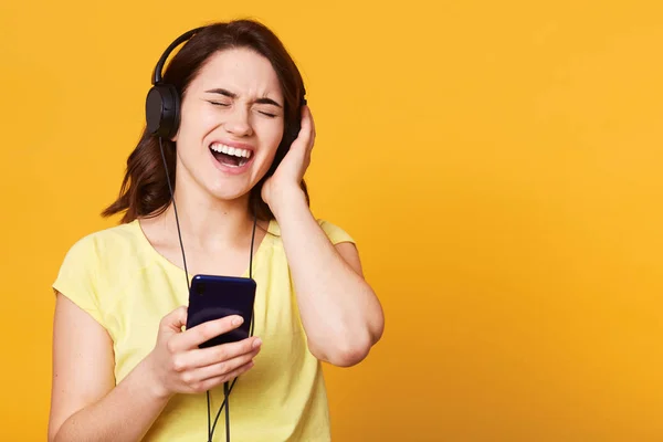 Happy beztroski młoda kobieta słuchania muzyki ze smartfona nad studiem tła, śpiewając piosenkę lubi głośno, stojący z zamkniętymi oczami, dotyka jej słuchawki, ubrany casual żółty t shirt. — Zdjęcie stockowe