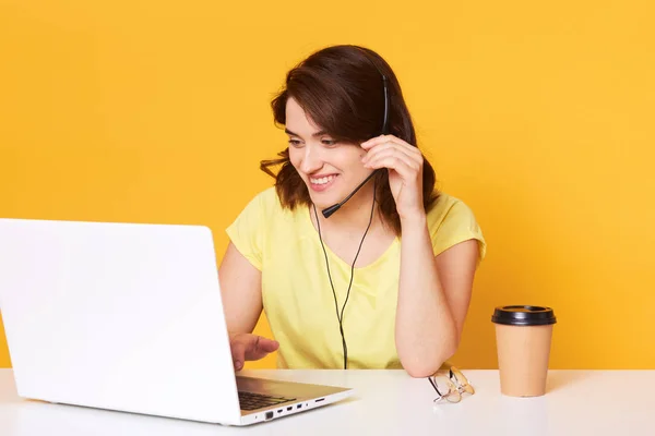 Gambar wanita berambut cokelat dengan kaos kasual kuning duduk di kursi di meja putih bekerja pada laptop di atas latar belakang studio, memiliki percakapan yang menyenangkan dengan klien, terus tangan pada garniture, bekerja secara online . — Stok Foto