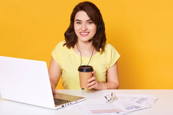 Όμορφη νεαρή γυναίκα κάθεται στο λευκό γραφείο μπροστά από το laptop και πίνοντας καφέ σε πακέτο από ένα Κύπελλο μίας χρήσης, θηλυκό φορώντας casual κίτρινο μπλουζάκι, δουλεύοντας σε απευθείας σύνδεση, θέτοντας απομονώνονται πάνω από το στούντιο τοίχου. — Φωτογραφία Αρχείου
