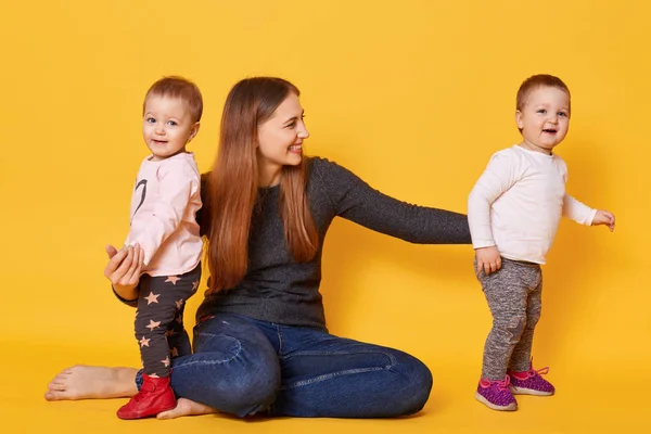 Image of attractive brown haired mother wants to be photographed with her sweet children, sit on floor in studio. Mother and girls twins, dressed casually, mom spends time with her doughters. — Stock Photo, Image