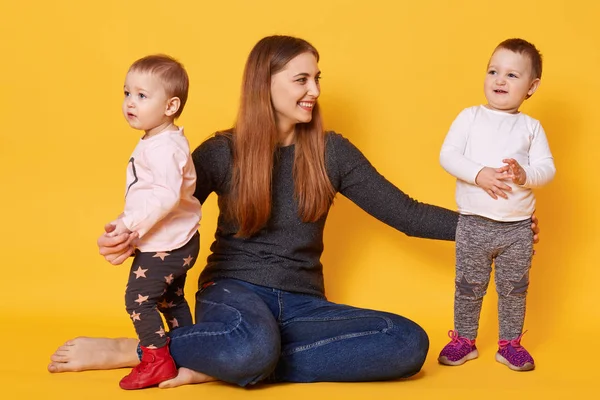 Happy woman, liitle twin girls, mother and her toddlers, try to make photo, infants play with mummy, poses in photo studio isoleted over yellow background. Motherhood, family, children concept. — Stock Photo, Image