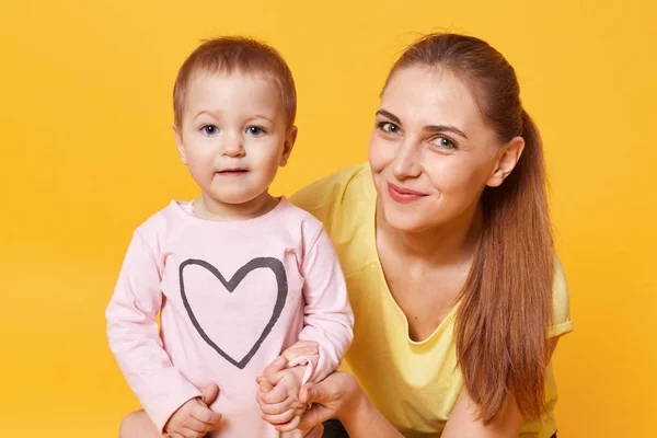 Close up portrait of beautiful young caucasian woman with her doughter, little girl wearing pink shirt with heart, mom dressed yellow t shirt, pose isolated over studio wall. Happy family concept. — Stock Photo, Image