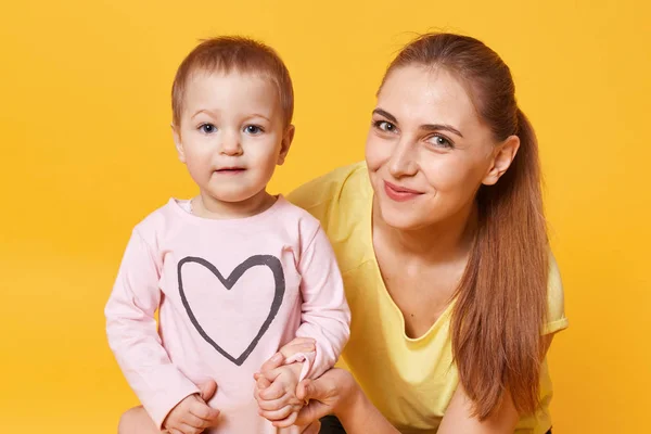 Potret wanita muda Kaukasia yang cantik dengan doughter-nya, gadis kecil mengenakan kemeja merah muda dengan hati, ibu berpakaian kuning t shirt, pose terisolasi di atas dinding studio. Konsep keluarga bahagia . — Stok Foto