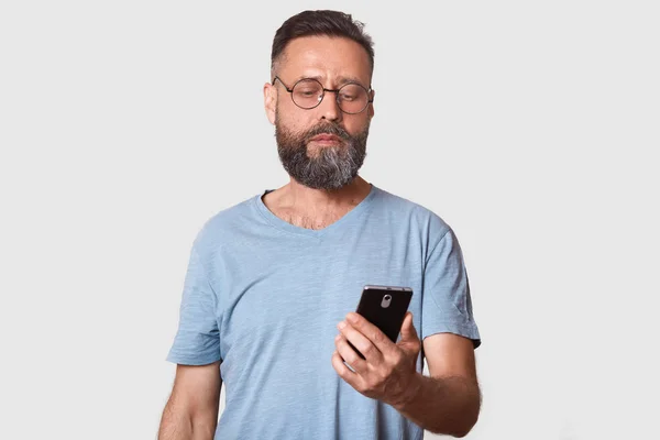 Schöner Mann mittleren Alters, der sein Telefon mit ernstem Gesichtsausdruck benutzt, während er vor grauem Hintergrund steht, attraktiver Mann, der wichtige Nachrichten von seiner Frau liest. Menschen und Technologie-Konzept. — Stockfoto