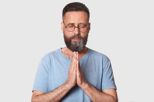 Studio shot of dark haired man in round spectacles, drerssed gray casual t shirt, holding hands together, praying for something, standing with closed eyes, against grey background. People concept. — Stock Photo, Image