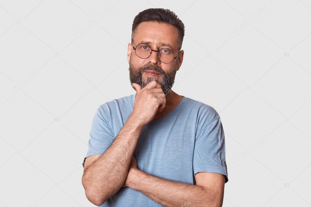 Portrait of middle bearded aged male with pensive facial expression, dressed gray cassual t shirt and round spectacles, keeps hand under chin, looks thoughtful, thinks abot new idea, has great plans.