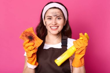 Hevesli gülümseyen ev hanımı kirli sünger ve temizlik deterjanı tutar, temizlik sonucu memnun, kirli olmak. Karizmatik esmer kadın kahverengi önlük, t shirt ve saç bandı giyer.