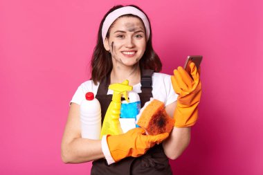 Beyaz rahat t shirt neşeli tatlı temizlik kadın portresi kadar yakın, kahverengi önlük ve saç bandı, sünger tutar, telefon ve temizlik deterjanları, pembe arka plan üzerinde izole, ev işi yapıyor.