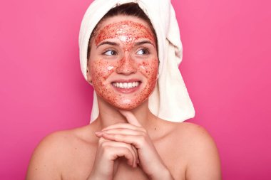 Gülümseyen samimi kadın bir kenara bakar, stüdyoda pembe arka plan üzerinde izole poz, çıplak olmak, yeni yüz maskesi çalışıyor, zevkle sonuç bekliyor, olumlu görünüyor. Güzellik ve cilt bakım konsepti.