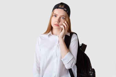 Uzun saçlı kalemli genç kız beyaz arka plan üzerinde duruyor, cep telefonu tutan, ciddi konuşma, dikkatle hoparlör dinleme, doğrudan kameraya bakar, rahat giysiler giyiyor.