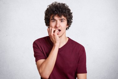 Koyu kıvırcık saçlı genç adamın stüdyo çekimi, rahat giyinmiş, tırnaklarını ısırma ve beyaz arka plan aleyhine dururken doğrudan kameraya bakarak, bir şey korkuyor. Kopyalama alanı.