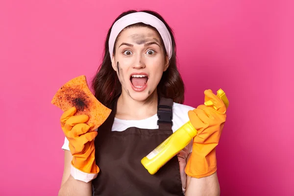 Impressed tired model posing isolated over pink background in studio, opening mouth with shock, doing household chores, wearing brown apron, t shirt and white headband. Housewife and cleaning concept. — Stock Photo, Image