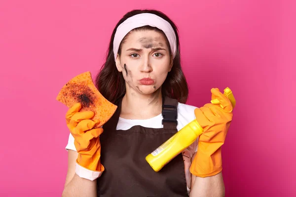 Disappointed exhausted black haired housewife has no energy to tidy up, looking directly at camera with upset facial expression, wearing white headband, brown apron and casual white t shirt. — Stock Photo, Image