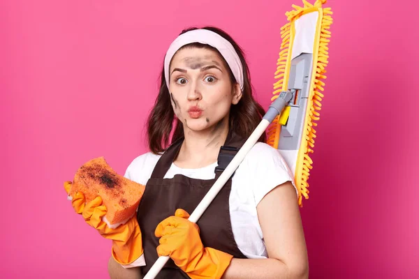 Indoor studio shot of beautiful hard working young housewife with wide opened eyes posing isolated over bright pink background holding special equipment in both hands, ready to do housework. — Stock Photo, Image