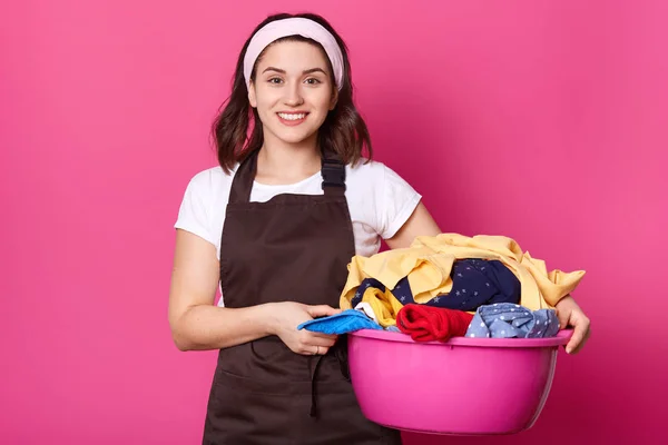 Young smiling beautiful female walking with pink basin full of dirty items of clothes, holding it with both hands, looks positive. Busy attractive housewife stands isolated over pink background. — Stock Photo, Image