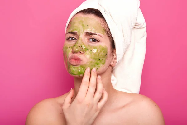Attractive emotional young beauty has green mask on her face, day to have spa procedures, protrudes her lips, has unpleasant facial expression. Magnetic model poses isolated over pink background. — Stock Photo, Image