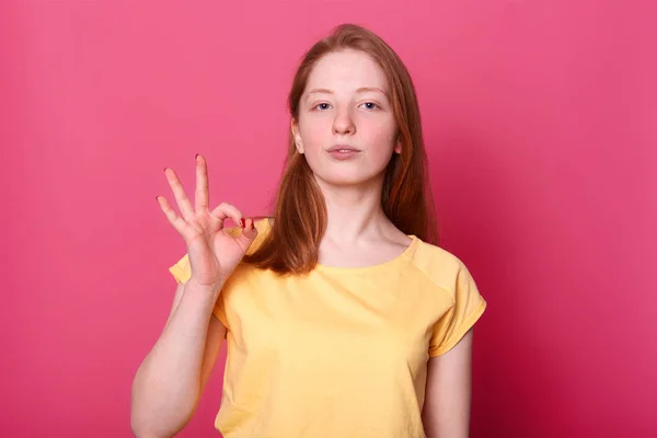 Close up studio ritratto di rosso dai capelli giovane ragazza in piedi con fermezza, mostrando segno positivo, sembra serio e sicuro di sé. Piccola ragazza affascinante con indosso maglietta gialla casual. Concetto di persone ed emozioni . — Foto Stock