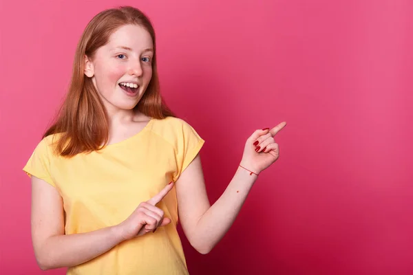 Retrato de linda encantadora encantadora adolescente alegre que muestra algo que apunta a un lado con los dedos delanteros, hembra está sonriendo sobre la pared del estudio. Espacio en blanco para texto promocional o publicidad . — Foto de Stock