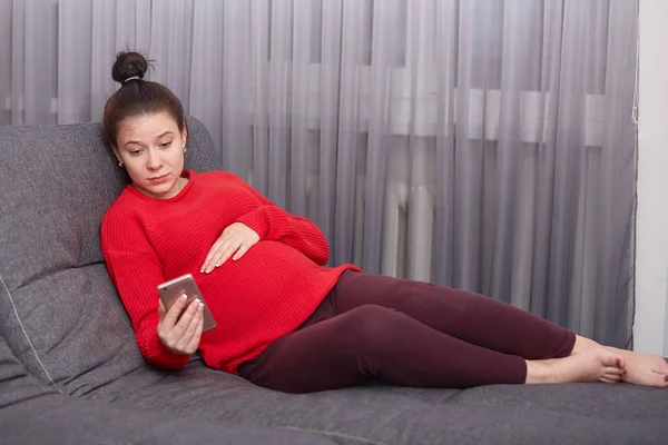 Uppmärksam allvarlig gravid ung dam med vila på soffan, hålla ena handen på magen, med telefon i handen, titta på video på nätet, tillbringar tid på mammaledighet, ser lugn och tankeväckande. — Stockfoto