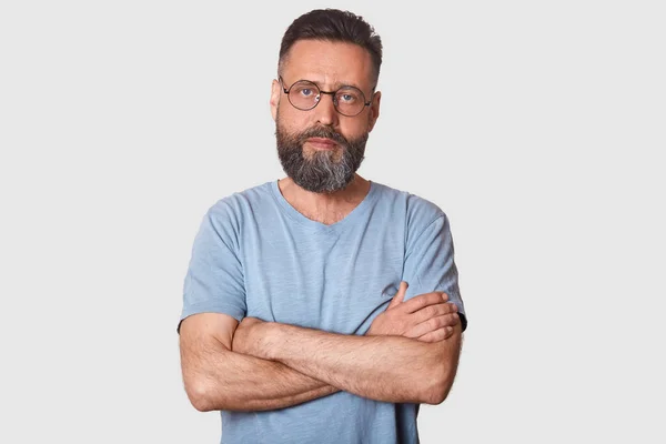 Serious magnetic black haired man posing with folded arms, having strong look, determined facial expression, having athletic arms. Middle aged bearded model poses isolated over light grey background.