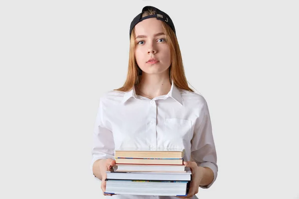 Blauäugige charmante Schulmädchen hat einen ernsten Gesichtsausdruck, steht gerade, hält Bündel verschiedener Bücher in beiden Händen, bereit, mit dem Lernen zu beginnen. Menschen und pädagogisches Konzept. — Stockfoto