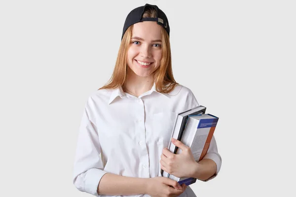 Die junge lächelnde Studentin posiert vor weißem Hintergrund mit einem Strauß bunter Bücher, sieht glücklich und zufrieden aus. blondes, aufrichtiges Model mit weißem Hemd, schwarzer Mütze. Menschen und Konzept studieren. — Stockfoto