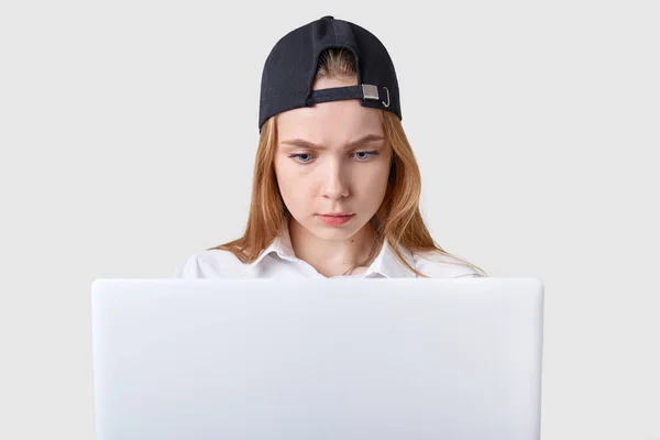 Chica seria reflexivo utiliza portátil blanco, busca nueva información, parece interesado en el tema, sentado aislado sobre fondo blanco. Joven modelo atractivo lleva blusa blanca y gorra negra . —  Fotos de Stock