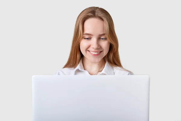 Sorrindo encantado menina de olhos azuis senta-se à mesa, trabalhando no laptop, rede de surf, lendo histórias engraçadas, wathing e ler posts em sites de redes sociais, parece feliz, vestindo blusa formal branca . — Fotografia de Stock