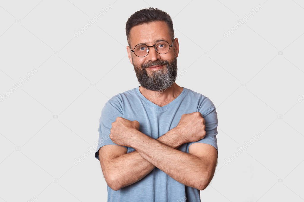 Kind pleasant good looking man with sincere look stands in front of camera, having confident look, holding his arms near chest, making cross, looks pleased and funny. People and emotions concept.