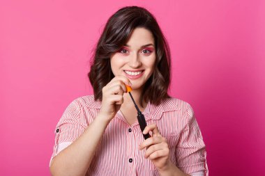 Kahverengi saçlı sevimli model portresi temiz taze deri ye sahiptir, çizgili gömlek giyen, pembe stüdyo arka planda poz ve kamera bakarak,, maskara tutan. Güzellik, makyaj, reklam konsepti.