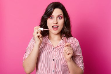 Horizontal studio portrait of beautiful young brunette woman in stiped blouse, holding open lip gloss near her face on pink background, has shoked facial expression, stands with opened mouth. clipart