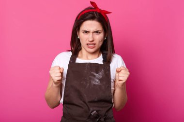 Görüntü kızgın kadın yumruklar içine ellerini sıktı, bayan tshirt, önlük, saç bandı giyen, pembe stüdyo duvarına karşı pozlar, kötü duygular vardır, kocası ile çatışma vardır. İnsanlar duygu kavramı.