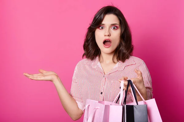 Plan studio de jeune femme caucasienne portant des sacs à provisions dans un geste surpris et excité, a les cheveux foncés, porte une chemise, achète dans un centre commercial, posant isolé sur fond rose avec espace de copie — Photo