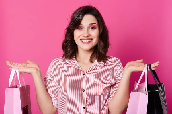 Close up retrato da bela mulher morena sorridente feliz com cabelo ondulado, camisa listrada vestida, detém sacos de compras. Modelo feminino fica sorrindo enquanto posando isolado sobre fundo de estúdio rosado . — Fotografia de Stock