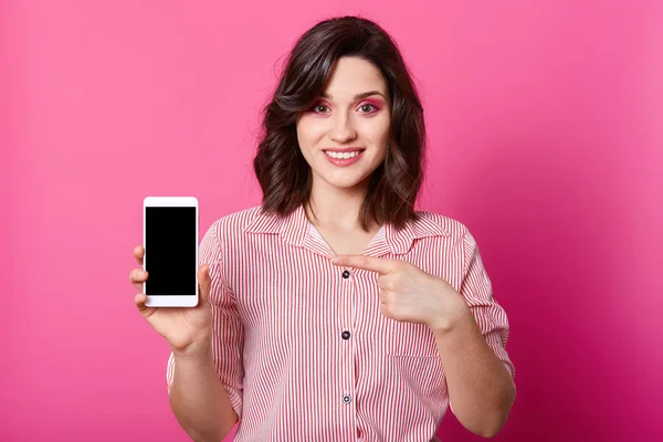 Студійний знімок привабливої жінки стоїть посміхаючись і вказуючи на мобільний телефон з порожнім екраном, відчуває себе щасливим, одягненим в елегантний одяг, моделі, що позують над рожевою стіною студії. Копіювати простір для просування . — стокове фото