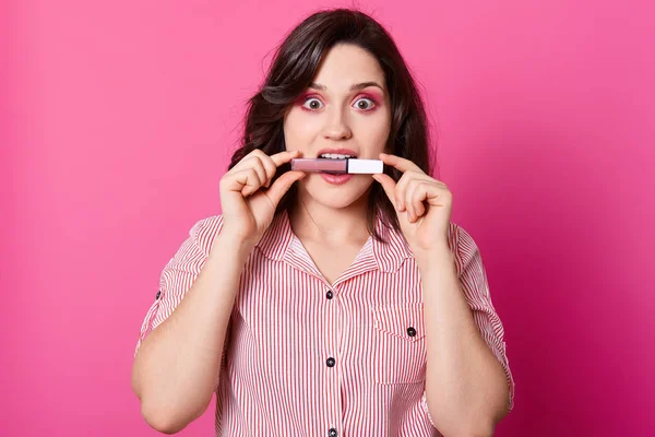 Hermosa mujer caucásica con el pelo negro ondulado, tiene maquillaje de moda, posando en el estudio con fondo rosa, sostiene brillo labial líquido con dientes, se levanta con expresión facial asombrada. Concepto de belleza . — Foto de Stock