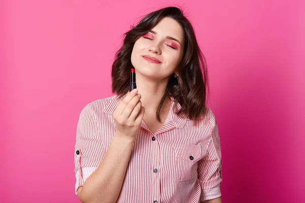 Foto de media longitud de la mujer está de pie con los ojos cerrados y la celebración de lápiz labial rojo. Retrato de belleza de modelo con cabello ondulado oscuro, blusa elegante vestida, posa en estudio con fondo rosa. Concepto de belleza . — Foto de Stock