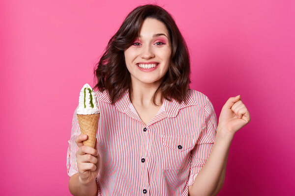 Studio shot of cheerful dark haired woman holds ice cream, clench fist, has happy facial expression, posing in striped shirt, has toothy smile, isolated on rose background. Happyness, emotions concept