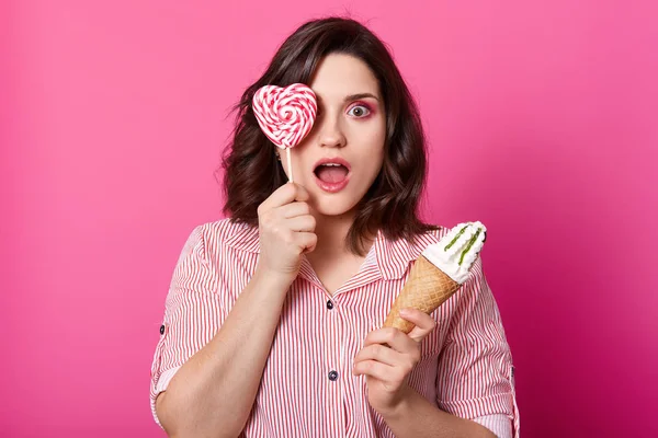 Surprised young woman with opened mouth looking camera with astonished facial expression, stands with ice cream in one hand and covering her eye with lolly pop, posing isolated over pink background.