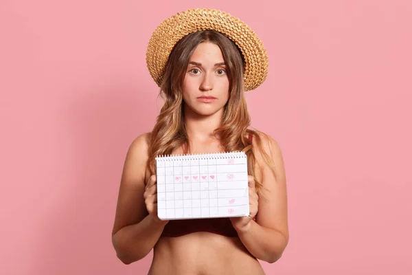 Horizontal shot of beautiful tanned young European female dressed straw hat and swimsuit, holding period calendar, has unhappy expression, has menstuation, isolated over pink studio background.